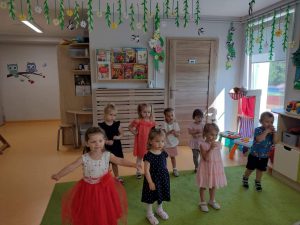 Dzieci stoją na zielonym dywanie ubrane odświętnie i tańczą do piosenki pt." Tato, Tato" . W tle widać sale zabaw.