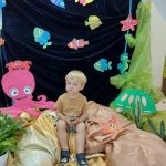 Zdjęcie przedstawia podwodny świat. Chłopczyk siedzi na jasno-brązowym materiale. Za nim widać niebieski materiał a na nim powieszone kolorowe rybki, ośmiornica i złów.