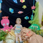 Zdjęcie przedstawia podwodny świat. Dziewczynka siedzi na jasno-brązowym materiale. Za nią widać niebieski materiał a na nim powieszone kolorowe rybki, ośmiornica i złów.