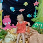 Zdjęcie przedstawia podwodny świat. Dziewczynka siedzi na jasno-brązowym materiale. Za nią widać niebieski materiał a na nim powieszone kolorowe rybki, ośmiornica i złów.