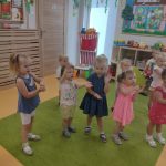 Dzieci stoją na zielonym dywanie i pokazują układ taneczny do piosenki o małym rekinie.