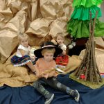 Dwie dziewczynki i chłopiec siedzą ubrani w góralskie stroje. Koło nich stoi drewniane palenisko. W tle widać dużą choinkę zrobioną z bibuły i góry wykonane z szarego papieru.
