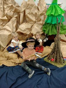 Dwie dziewczynki i chłopiec siedzą ubrani w góralskie stroje. Koło nich stoi drewniane palenisko. W tle widać dużą choinkę zrobioną z bibuły i góry wykonane z szarego papieru.