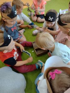 Dzieci siedzą na zielonym dywanie. Na głowie mają założone opaski w kształcie pirackiej czapki. Dzieci oglądają biżuterię znajdującą się w drewnianej brązowej skrzynce.