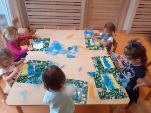 Dzieci siedzą przy stoliku i na żółtej kartce przyklejają niebieską folię.