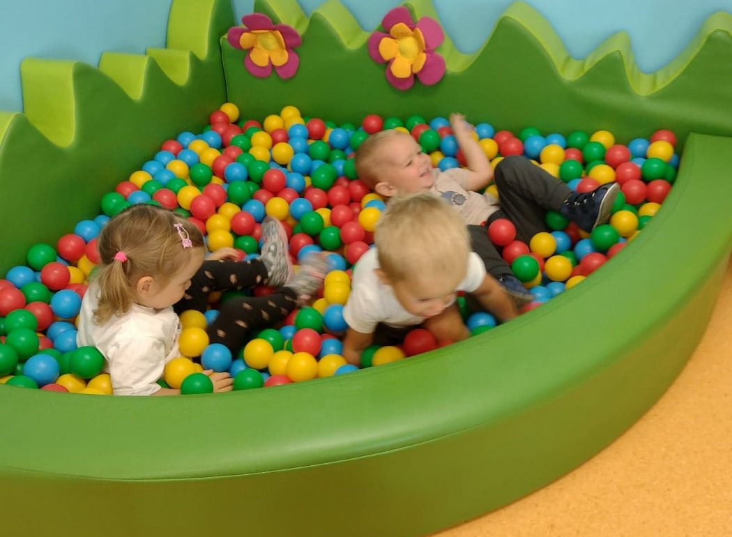 Dzieci siedzą w zielonym basenie z kolorowymi kulkami i rzucają nimi do siebie.