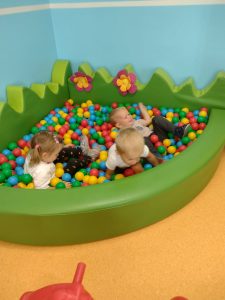Dzieci siedzą w zielonym basenie z kolorowymi kulkami i rzucają nimi do siebie.