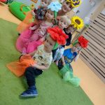 Dzieci przebrane w kolorowe kwiaty z opaskami na głowie siedzą na zielonym dywanie z opiekunką i pozują do zdjęcia.