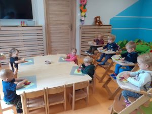 Dzieci siedzą przy stoliku i w drewnianych krzesełkach, przed sobą mają szarą podkładkę na której leży zielona kartka z narysowanym wazonem. Dzieci przyklejają wydrukowane kwiaty tworząc jesienny bukiet.