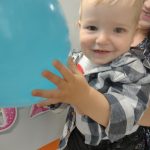 Opiekunka trzyma na rękach chłopca. W rączkach trzyma niebieskiego balonika i uśmiecha się do zdjęcia.