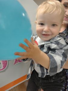 Opiekunka trzyma na rękach chłopca. W rączkach trzyma niebieskiego balonika i uśmiecha się do zdjęcia.