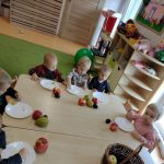 Dzieci siedzą przy stoliku, przed sobą mają położony biały talerzyk i owoce. Dzieci degustują pokrojone owoce- jabłko, gruszkę i śliwkę.