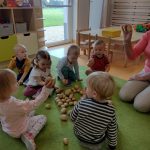 Dzieci siedzą razem z opiekunką na zielonym dywanie i oglądają rozsypane ziemniaki. W tle widać sale zabaw.