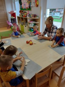 Dzieci siedzą przy stole w niebieskich fartuszkach i lepią jabłka z masy solnej.