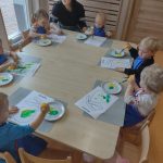 Dzieci siedzą przy stoliku w niebieskich fartuszkach i malują na białej kartce sylwetkę gruszki zieloną i żółtą farbą.