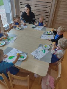 Dzieci siedzą przy stoliku w niebieskich fartuszkach i malują na białej kartce sylwetkę gruszki zieloną i żółtą farbą.