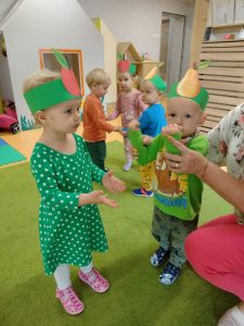 Dziewczynka z chłopcem ubrana na zielono z opaskami na głowie w kształcie jabłka i gruszki, klaszczą w dłonie w rytm piosenki. W tle widać troje dzieci trzymające się za rączki i tańczące w kółeczku.