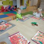 Dzieci siedzą przy stoliku i przyklejają na kartce wymalowanej czerwoną i żółtą farbą, brązowo- białego grzybka. W tle witać sale zabaw.