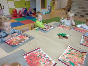 Dzieci siedzą przy stoliku i przyklejają na kartce wymalowanej czerwoną i żółtą farbą, brązowo- białego grzybka. W tle witać sale zabaw.
