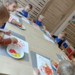 Dzieci siedzą przy stoliku ubrane w niebieskie fartuszki i malują czerwoną i żółtą farbą paluszkami po białej kartce.