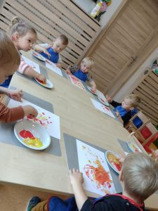 Dzieci siedzą przy stoliku ubrane w niebieskie fartuszki i malują czerwoną i żółtą farbą paluszkami po białej kartce.