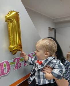 Opiekunka trzyma chłopca na rękach. Chłopiec dłonią wskazuje na dużą złotą cyfrę jeden, wiszącą nad urodzinowym napisem.