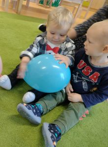 Chłopcy siedzą na zielonym dywanie i trzymają niebieski balonik.