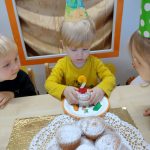 Chłopczyk siedzi przy stole w urodzinowej czapce, w dłoniach trzyma babeczkę ze świeczką w kształcie cyfry dwa.