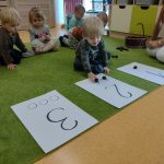 Dzieci siedzą na zielonym dywanie przed sobą mają rozłożone trzy kartki z cyframi. Dzieci mają za zadanie ułożyć tyle kasztanów na kartce jaką widzą cyfrę na kartce przed sobą..
