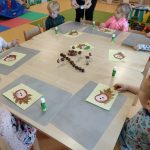 Dzieci siedzą przy stoliku i na jasno-żółtej kartce przyklejają wysuszonego liścia a na nim kasztanka z papieru.