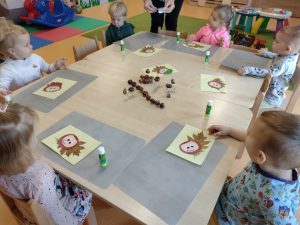 Dzieci siedzą przy stoliku i na jasno-żółtej kartce przyklejają wysuszonego liścia a na nim kasztanka z papieru.