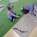 Dzieci siedzą na dywanie i układają z kasztanów wzory narysowane na dużym szarym papierze.