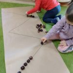 Dzieci siedzą na dywanie i układają z kasztanów wzory narysowane na dużym szarym papierze.