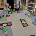 Dzieci siedzą przy stoliku i na czarnym prostokącie przyklejają trzy kółka w kolorze czerwonym, żółtym i zielonym, tworząc z nich sygnalizator świetlny.