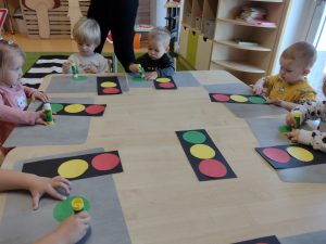 Dzieci siedzą przy stoliku i na czarnym prostokącie przyklejają trzy kółka w kolorze czerwonym, żółtym i zielonym, tworząc z nich sygnalizator świetlny.