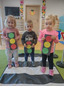 Troje dzieci stoją na rozłożonej macie emitującej przejście dla pieszych. W dłoniach trzymają zrobione przez siebie z papieru kolorowego sygnalizatory świetlne.