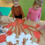 Dzieci stoją przy stoliku i przyklejają czerwone liście na gałązkach namalowanych na szarym papierze.