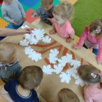 Dzieci stoją przy stoliku i przyklejają białe liście na gałązkach namalowanych na szarym papierze.