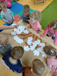 Dzieci stoją przy stoliku i przyklejają białe liście na gałązkach namalowanych na szarym papierze.