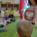 Dzieci siedzą po turecku na zielonym dywanie i oglądają flagę i godło Polski trzymane w dłoniach przez opiekunkę.