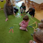 Dzieci razem z opiekunką siedzą na dywanie i układają z kolorowych papierowych kredek różne konstrukcje np. domek, płotek, drzewko.