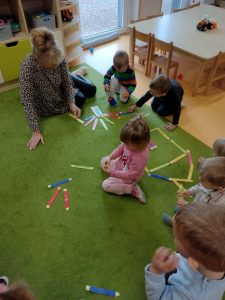 Dzieci razem z opiekunką siedzą na dywanie i układają z kolorowych papierowych kredek różne konstrukcje np. domek, płotek, drzewko.