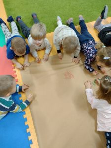Dzieci leżą na brzuszku przed rozłożonym dużym szarym papierze i rysują różnymi rodzajami kredek.