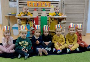Dzieci siedzą po turecku koło siebie i w drewnianych krzesełkach. Na głowie mają ubrane opaski przedstawiające kolorowe kredki. W tle widać kolorowe kredki zrobione z kartonu i napis 'Kredki".