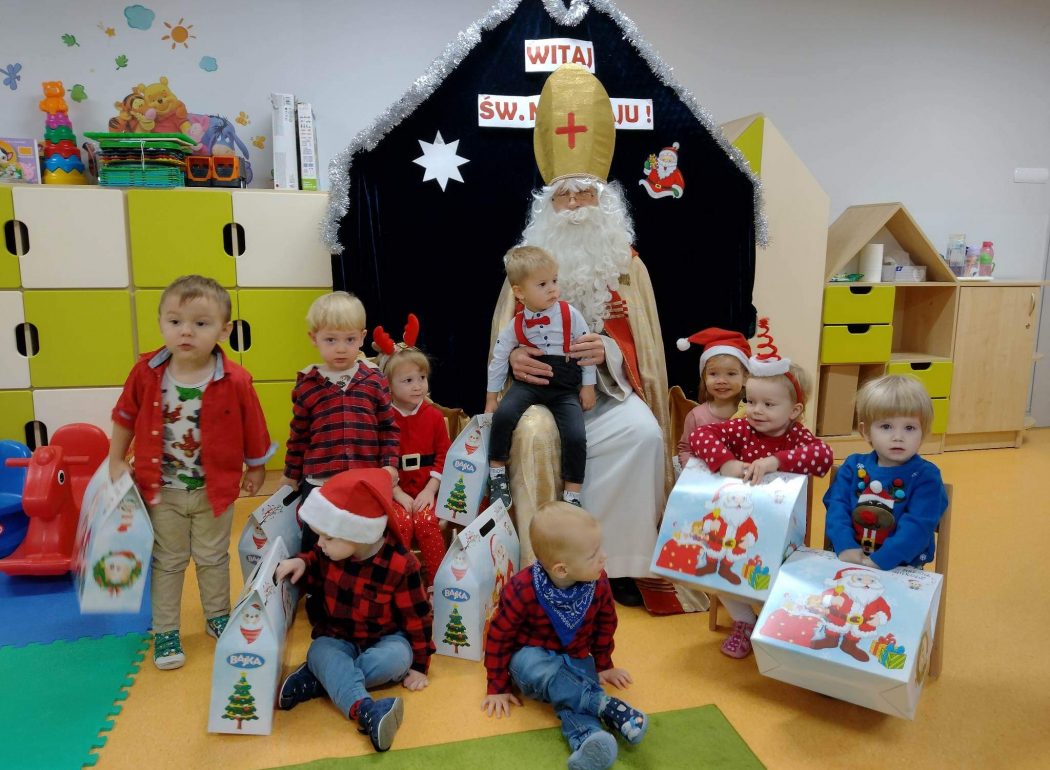 Dzieci siedzą z prezentami koło Św. Mikołaja i pozują do wspólnego zdjęcia. W tle widać napis "Witaj Św. Mikołaju!" na ciemno granatowym materiale.