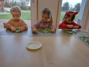 Dzieci siedzą przy stoliku i na szarej podkładce ozdabiają kolorowymi kółeczkami papierowy zielony wianuszek.