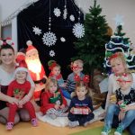 Dzieci wraz z opiekunkami ubrane świątecznie pozują do zdjęcia w scenerii świąteczno-zimowej.