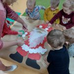 Dzieci stają przy stoliku i przyklejają razem z opiekunką białe okrągłe waciki na namalowanej sylwetce Św. Mikołaja.