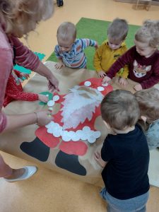 Dzieci stają przy stoliku i przyklejają razem z opiekunką białe okrągłe waciki na namalowanej sylwetce Św. Mikołaja.