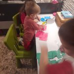 Dzieci siedzą przy stoliku i przyklejają kolorowe karteczki tworząc z nich łańcuch na choinkę.